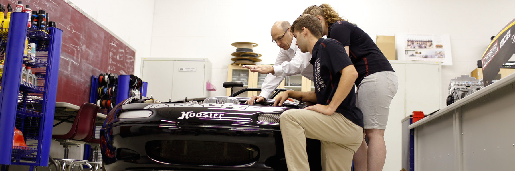 An instructor and two students examining the engine of a race car.